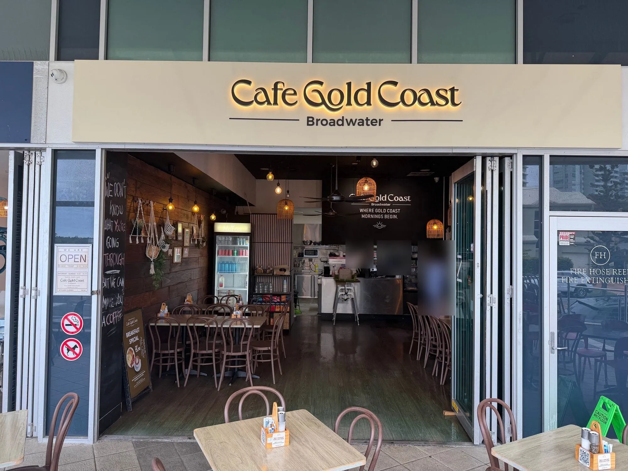 Cafe Gold Coast at Aqua, its cream sign reading "Cafe Gold Coast, Broadwater" above folding doors opened to the walkway.