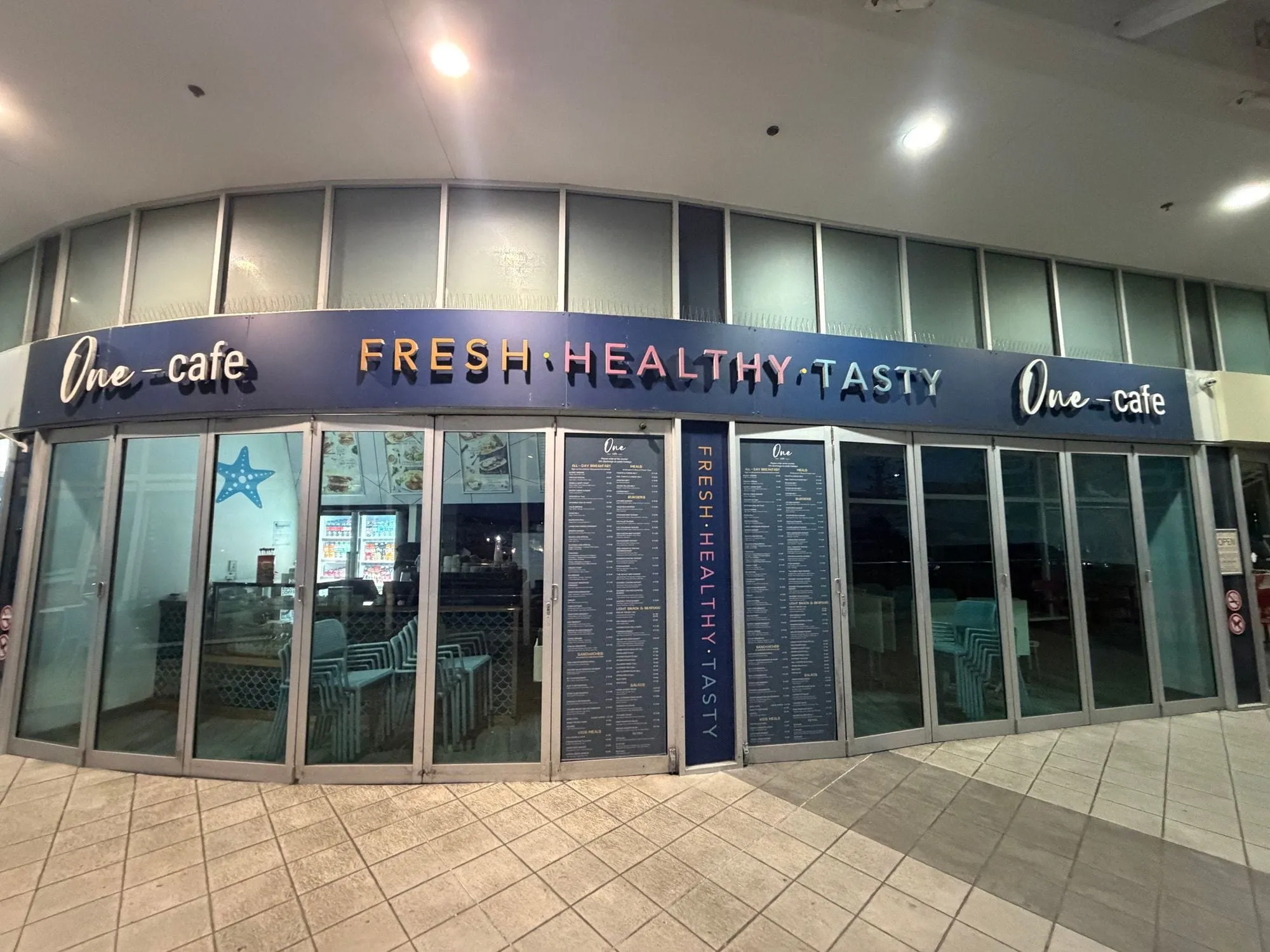 One-cafe at Aqua, its frontage lettered "fresh, healthy, tasty" along the glass, lit from inside in the evening.