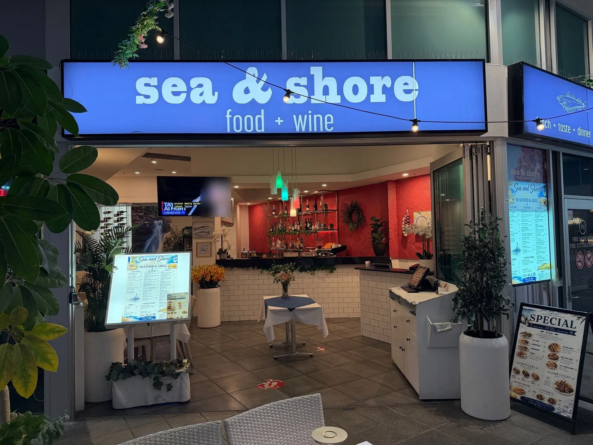The Sea & Shore shopfront at Aqua, a blue illuminated sign reading "sea & shore, food + wine" above the open frontage.