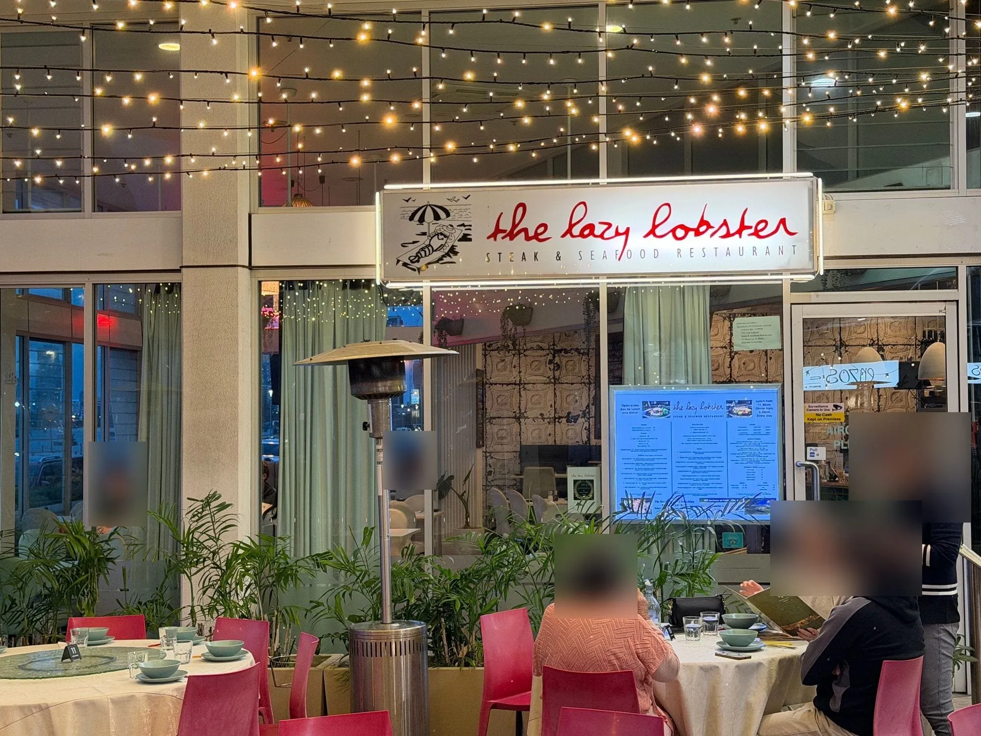The Lazy Lobster at Aqua — a hanging sign reading "the lazy lobster, steak & seafood restaurant" above covered outdoor tables strung with festoon lights.