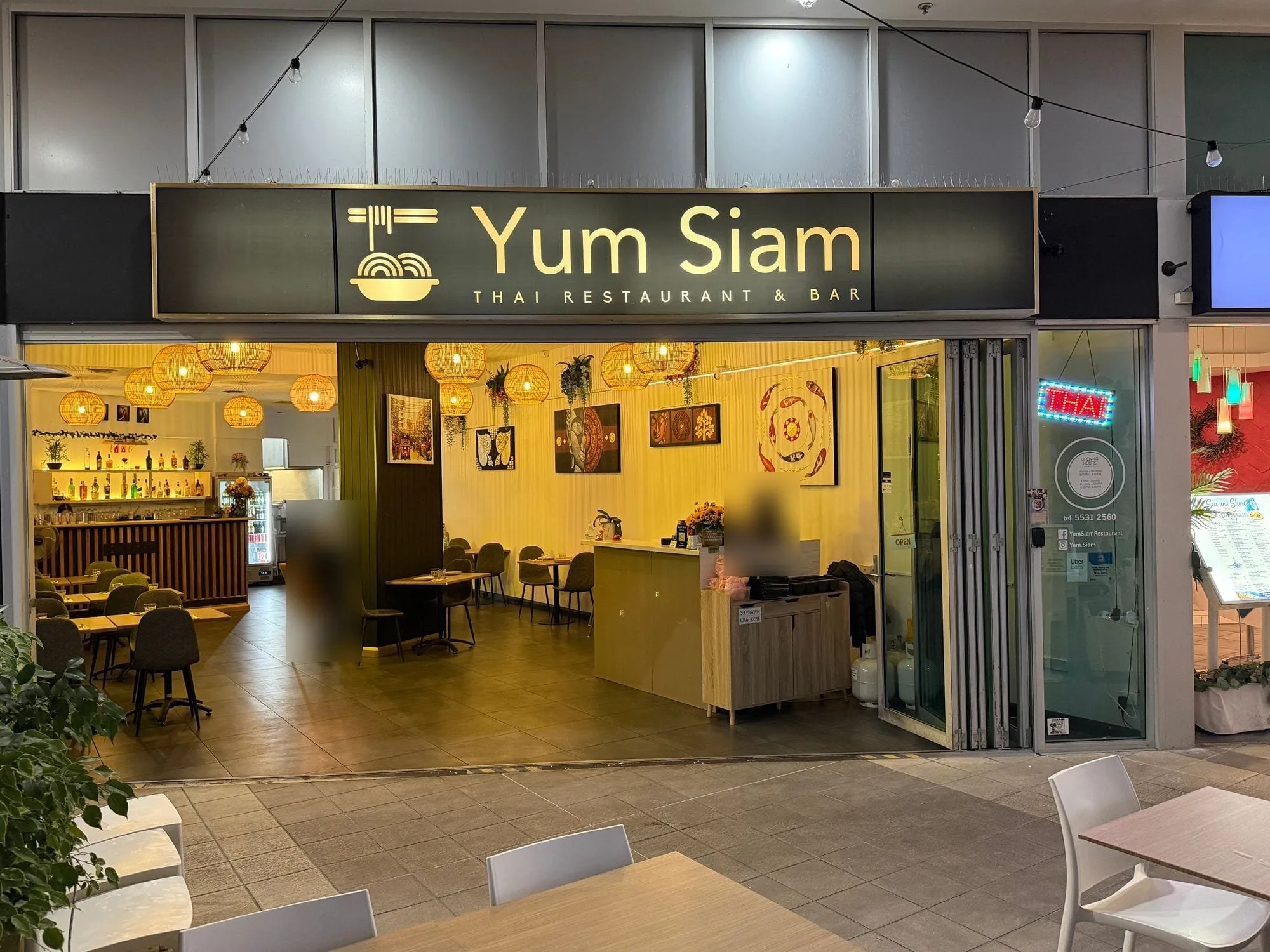 Yum Siam at Aqua, a dark sign with a gold noodle-bowl emblem reading "Yum Siam, Thai Restaurant & Bar", with a warmly lit dining room behind.