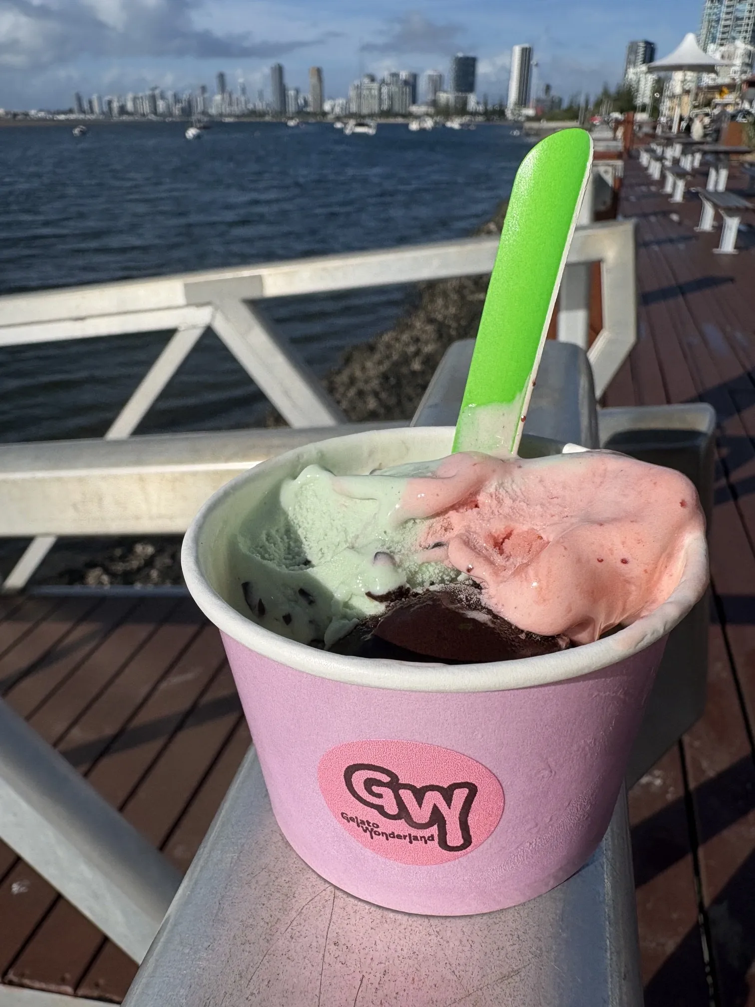 A Gelato Wonderland cup of three scoops with a spoon in it, resting on a timber jetty rail above the Broadwater.